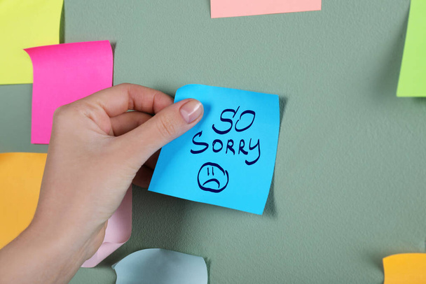 Apology. Woman sticking paper note with text So Sorry and drawn sad face onto grey wall, closeup - Photo, Image