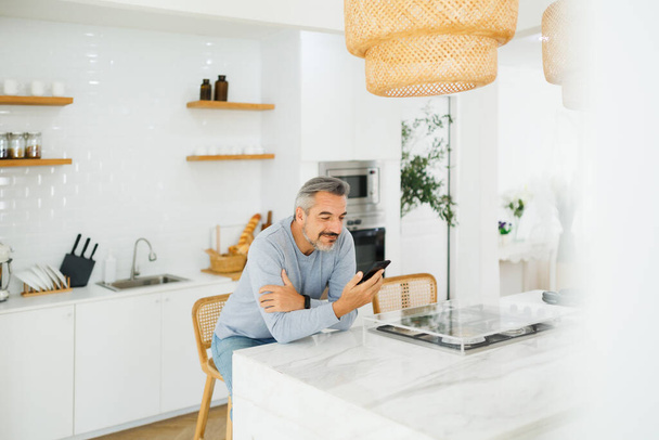 Happy Mature adult male using smartphone or mobile phone at home. Middle aged Man texting and reading message on the phone in kitchen. - Φωτογραφία, εικόνα