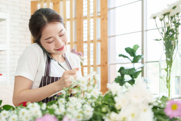 Smiling Asian Chinese florist woman calling on smartphone and taking notes in notebook at flower shop, Female receiving order of flowers, writing - Фото, изображение