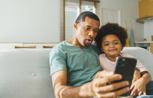 Smiling African American father and his cute boy using smartphone at home, African family doing video call on mobile phone, Black Dad and son sitting on sofa taking selfies. - Photo, image