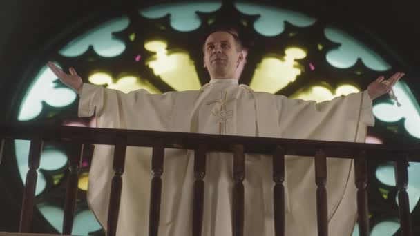 Low angle slowmo van priester in lange witte jurk prediking preek en kruising, staande tegen mooie glas-in-lood raam in de katholieke kerk - Video