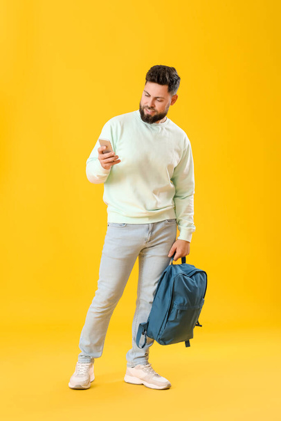 Sırt çantalı genç sakallı adam sarı arka planda cep telefonu kullanıyor. - Fotoğraf, Görsel