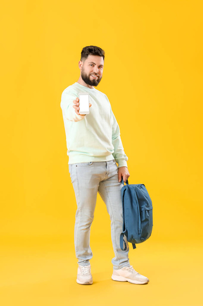 Young bearded man with backpack and mobile phone on yellow background - Foto, imagen