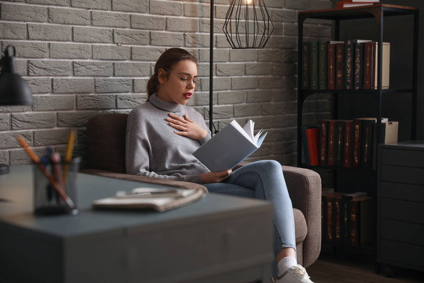 Evdeki koltukta kitap okuyan endişeli genç bir kadın. - Fotoğraf, Görsel