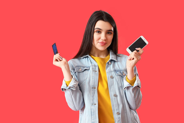 Kaunis nuori nainen matkapuhelimen ja luottokortin kanssa punaisella taustalla - Valokuva, kuva
