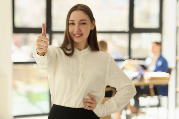 Cheerful young woman in formal outfit shows thumb up smiling and looking in camera. Female employee stands in office against colleagues at table - Foto, Imagem
