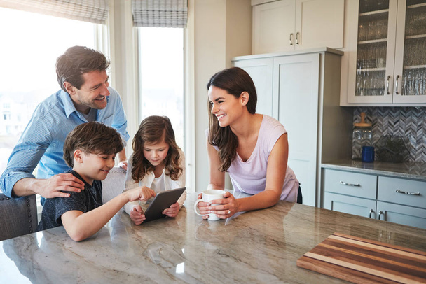 Technology keeps them close. a married couple and their young children playing with a tablet together in their kitchen - 写真・画像