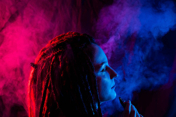 Profile of caucasian girl with dreadlocks smokes a vape in red blue light - Valokuva, kuva