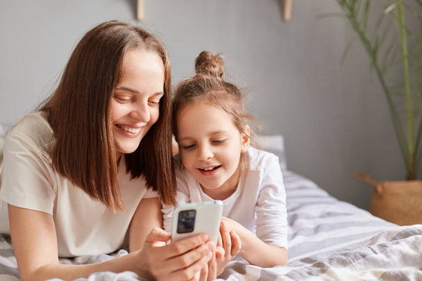 Happy mother and cute daughter using mobile phone, resting on bed at home, little girl with mommy looking at cell phone display watching video, shopping online. - Фото, зображення