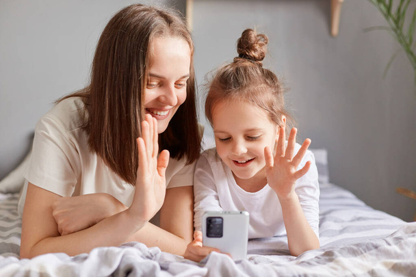 Joyful cheerful mother and daughter using mobile phone, relaxing on bed at home looking at smartphone screen having video call, waving hand saying hello. - Photo, image