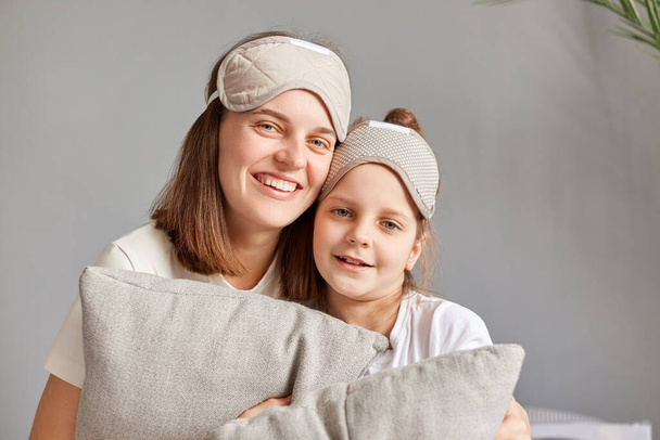 Cheerful mother with her cute little daughter sitting on bed, enjoying together in free time hugging each other holding pillows looking at camera with smile. - Foto, Bild