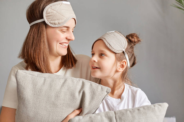 Smiling woman wearing sleeping mask sitting at home with her cute little daughter on bed talking looking at each other expressing positive emotions. - Photo, Image