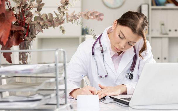 Skillful woman physician sitting at table in her office and writing. - Photo, Image