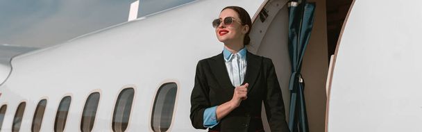 Woman flight attendant in sunglasses standing on airplane stairs at airport. Blurred background - Foto, Imagem
