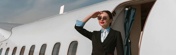 Улыбающаяся стюардесса в солнечных очках, стоящая на лестнице самолета в аэропорту - Фото, изображение