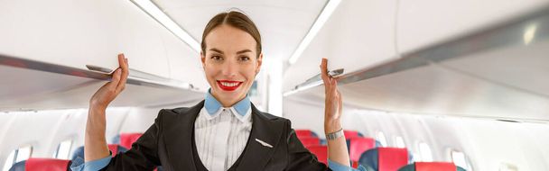 Joyful woman flight attendant looking at camera and smiling while standing between passenger chairs in aircraft - Foto, Bild