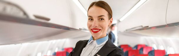 Cheerful woman stewardess or air hostess looking at camera and smiling while placing hand on passenger chair in aircraft - Valokuva, kuva