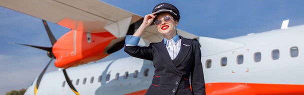 Fröhliche Flugbegleiterin in Airline-Uniform berührt Mütze und lächelt, während sie in der Nähe von Flugzeugen auf dem Flugplatz steht - Foto, Bild