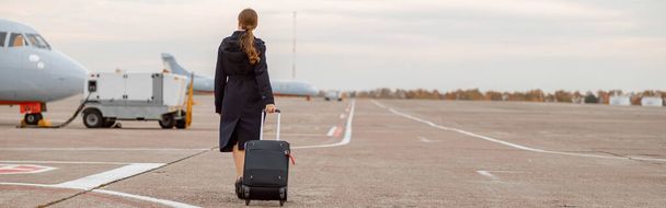 Back view of female traveler in black coat strolling down airport and carrying trolley luggage bag - Fotoğraf, Görsel