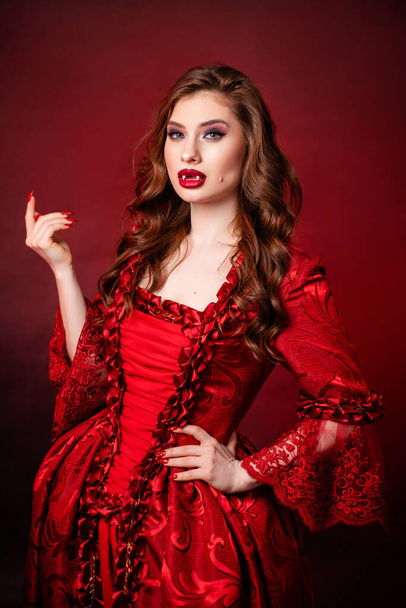Portrait of a young, attractive vampire woman in a red rococo dress posing isolated against a dark background with red backlights. - 写真・画像