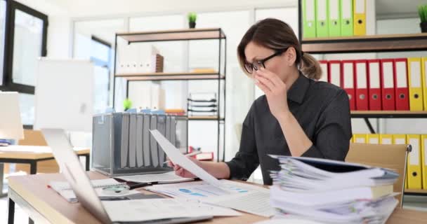 疲れた女社員がノートパソコンで机に座っている紙を見て眼鏡を外します。オフィスワーカーが職場で業務データを確認するのを心配 - 映像、動画