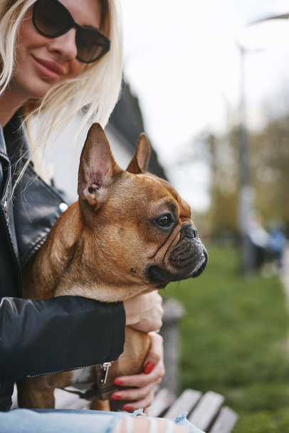 Young blonde woman sitting with a French bulldog puppy on laps on a bench in a park. Portrait of loving pet owner hugging a dog - Фото, зображення