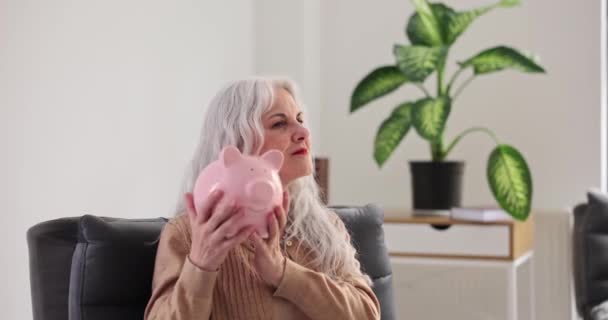 Hymyilevä kypsä nainen ravistelee säästöpossua kolikoilla olohuoneessa. Positiivinen iäkäs nainen tarkistaa rahaa säästöjä kotona. Rahoitusvarat - Materiaali, video