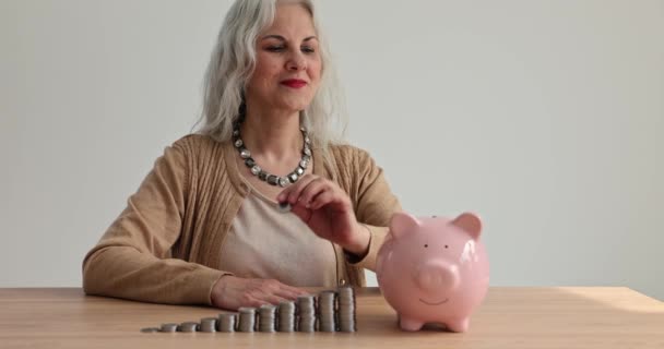 Happy senior woman puts coin into piggy bank at wooden table with cash money coins stacks slow motion. Save pension fund for financial savings concept - Footage, Video