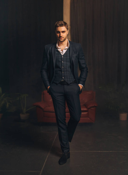 Portrait Handsome brutal man in classic strict three-piece suit jacket, trousers white shirt dark blue checkered vest. Successful businessman sexy guy stubble on face looks at camera. dark black room. - Foto, Bild