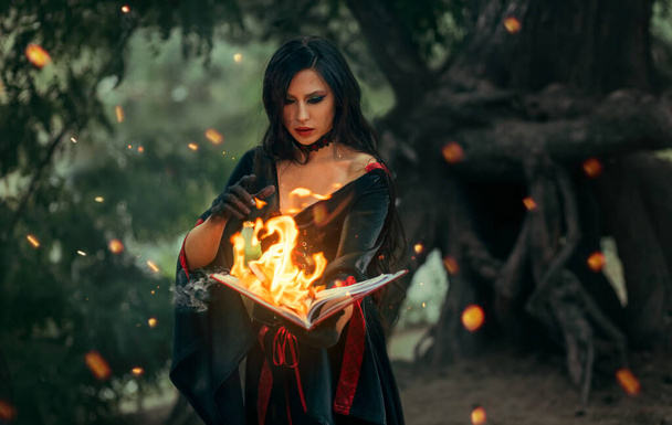 Fantasy halloween woman witch holds old burning magic book in hand, reads spell Paper page in bright flame fire light. Gothic girl sexy face in ashes, black dress costume. Dark night forest trees. Art - Photo, image