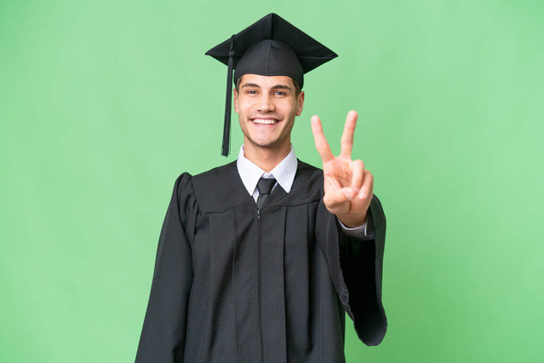Νεαρός απόφοιτος πανεπιστημίου καυκάσιος άνδρας πάνω από απομονωμένη φόντο χαμογελώντας και δείχνοντας σημάδι νίκης - Φωτογραφία, εικόνα