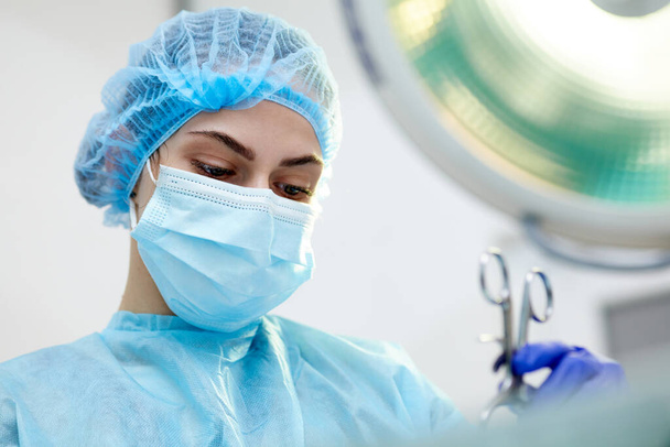 Close-up image of young woman, doctor, surgeon in protective medical mask leading surgery in medical clinic, hospital in surgery room. Concept of medicine, hospital, healthcare, treatment, profession - Photo, image