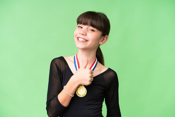 Маленькая кавказская девочка с медалями на изолированном фоне, показывающая большой палец вверх - Фото, изображение