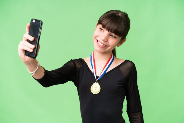 Pequeña chica caucásica con medallas sobre fondo aislado haciendo una selfie - Foto, Imagen