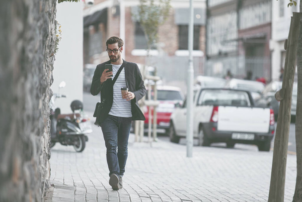 Auf seinem Weg durch die Stadt. ein stylischer junger Mann, der ein Handy benutzt, während er in der Stadt spaziert - Foto, Bild