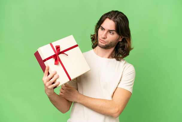 Νεαρός όμορφος άντρας που κρατάει ένα δώρο πάνω σε απομονωμένο φόντο με θλιβερή έκφραση - Φωτογραφία, εικόνα