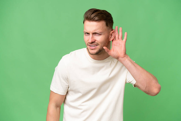 Jonge blonde blanke man over geïsoleerde achtergrond luisteren naar iets door het leggen van de hand op het oor - Foto, afbeelding