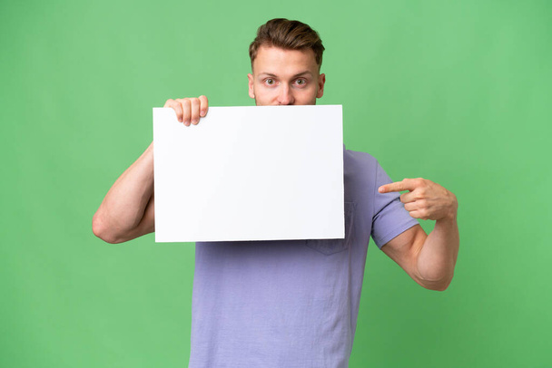 Νεαρός ξανθός καυκάσιος άντρας πάνω από απομονωμένο φόντο που κρατάει ένα άδειο πλακάτ και κρύβεται πίσω από αυτό - Φωτογραφία, εικόνα