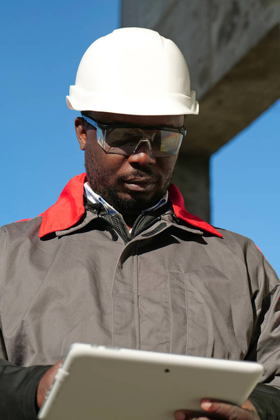 Αφροαμερικανός εργαζόμενος με σκληρό καπέλο και προστατευτικά γυαλιά με tablet υπολογιστή στέκεται στο εργοτάξιο - Φωτογραφία, εικόνα