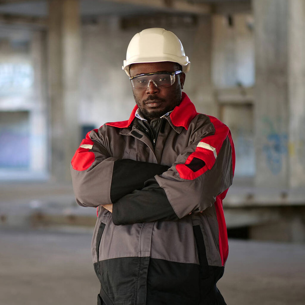 Africký americký pracovník v bílém tvrdém klobouku a ochranných brýlích stojí na staveništi a dívá se do kamery - Fotografie, Obrázek