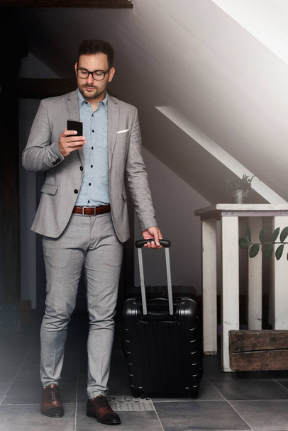 Elegant businessman with luggage in his hotel using smartphone.Traveling for work. - 写真・画像