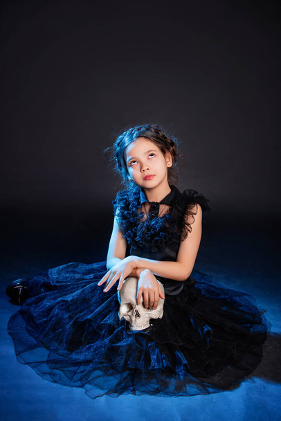A little girl in a black dress with a pigtail hairstyle on her head poses sitting with a skull in her hands, isolated on a dark background with blue backlight. - 写真・画像