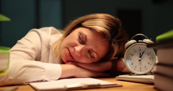 Mujer cansada se duerme en la mesa cerca de reloj despertador vintage y portapapeles con notas. Un empleado soñoliento cierra los ojos y se queda hasta tarde en la oficina - Imágenes, Vídeo