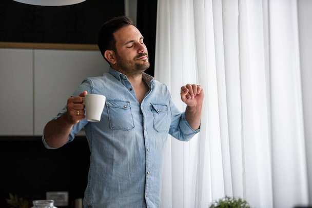 chico guapo está bebiendo café, de pie cerca de la ventana  - Foto, Imagen