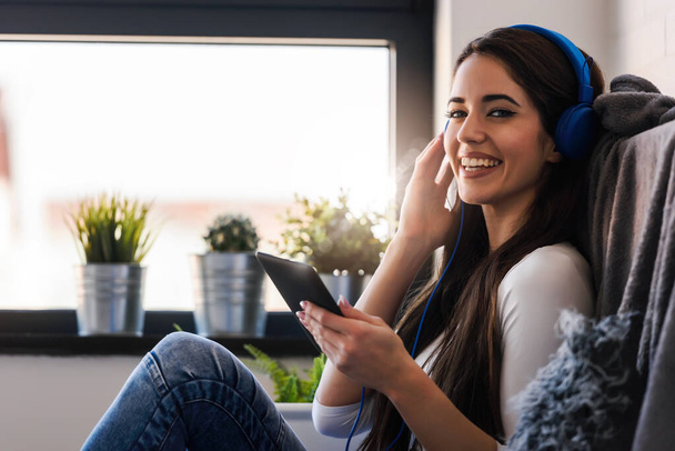 junge Frau mit Kopfhörern auf Sofa sitzend und Musik auf Tablet hörend - Foto, Bild