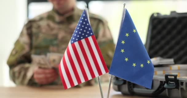 Drapeaux des États-Unis et de l'Union européenne contre les officiers militaires comptant l'argent comme aide internationale. L'homme en uniforme militaire est assis à table au bureau au ralenti - Séquence, vidéo
