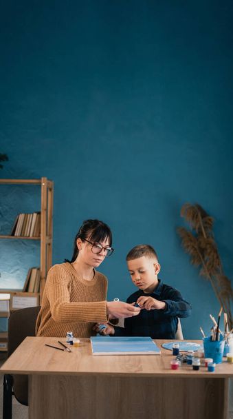 Een jonge mooie vrouw leert haar schoolzoon thuis te schilderen met olieverf op doek. Een vrouw brengt tijd door met haar kind. Een spandoek. kopieerruimte. - Foto, afbeelding