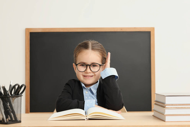 Happy little school child raising hand while sitting at desk with books near chalkboard - Photo, Image