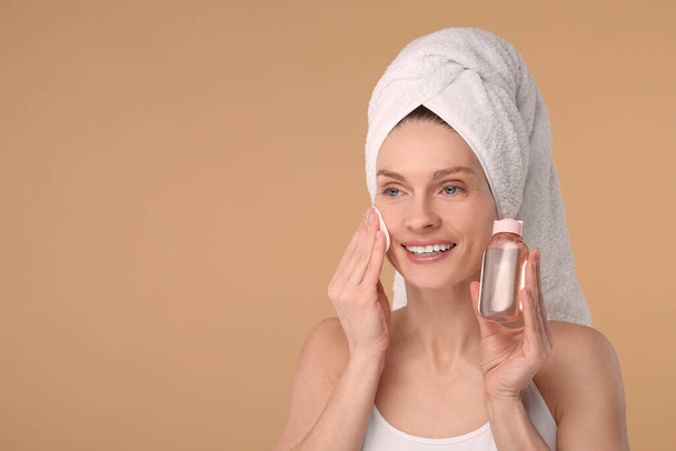 Beautiful woman in terry towel removing makeup with cotton pad on beige background, space for text - Фото, зображення