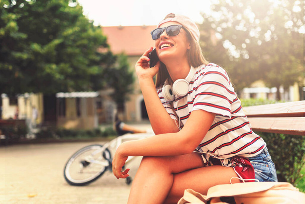 Chica sonriente con gafas de sol sentado en un banco en el verano utilizando el teléfono inteligente - Foto, Imagen